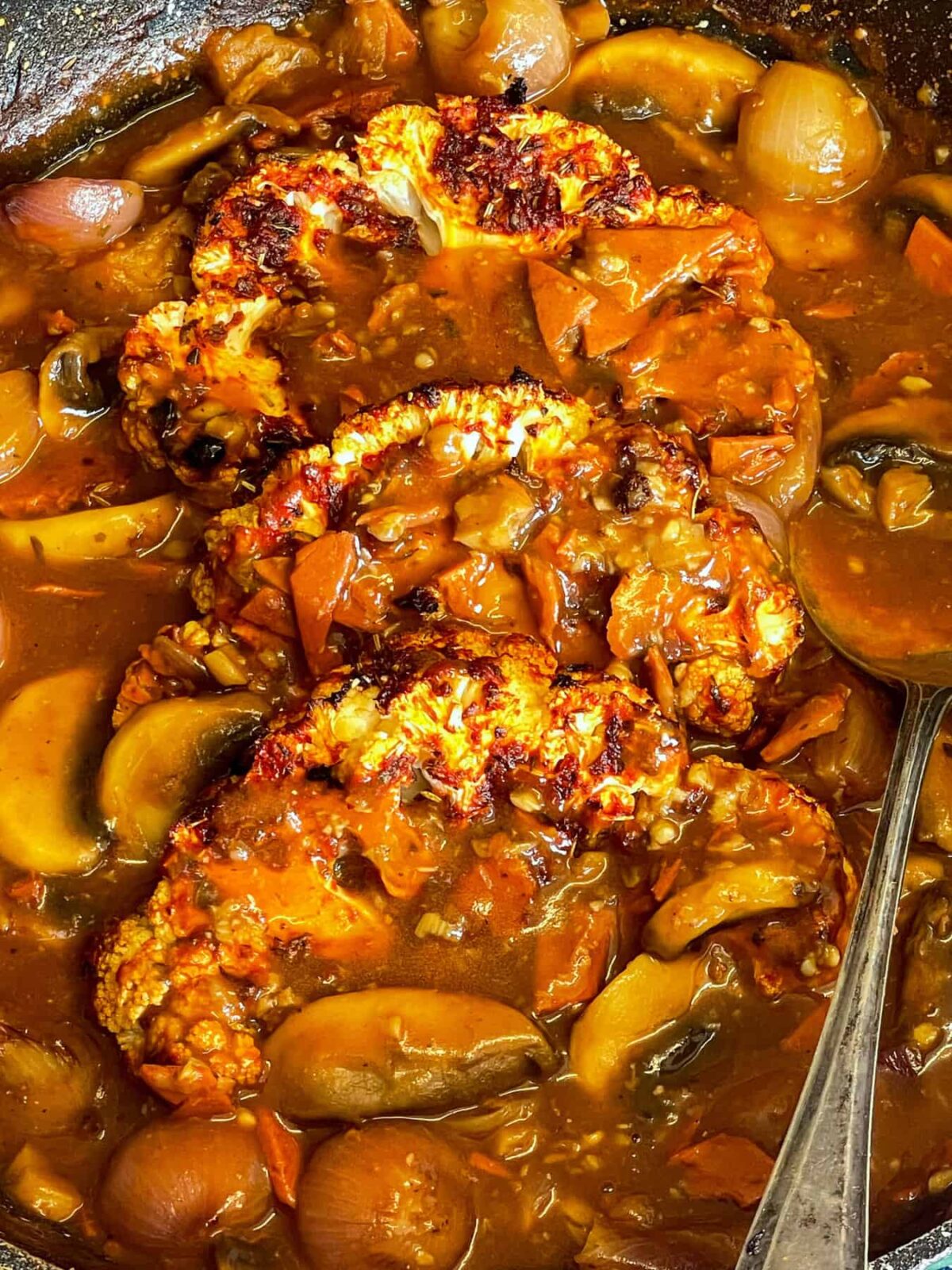 Cauliflower steaks added to pan with coq au vin sauce, silver serving spoon to side.