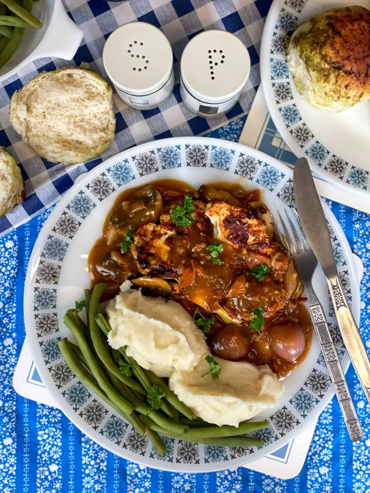 A blue and white patterned plate with vegan coq au vin served with mashed potatoes and green beans, bread rolls to side with salt and pepper shakers.