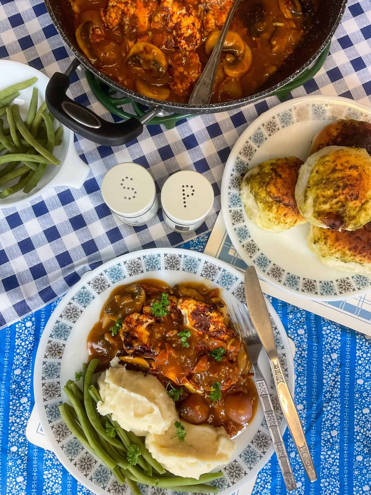 Served coq au vin with potatoes and green beans, small bread rolls to side, pan of coq au vin in background, blue and white check tablecloth.