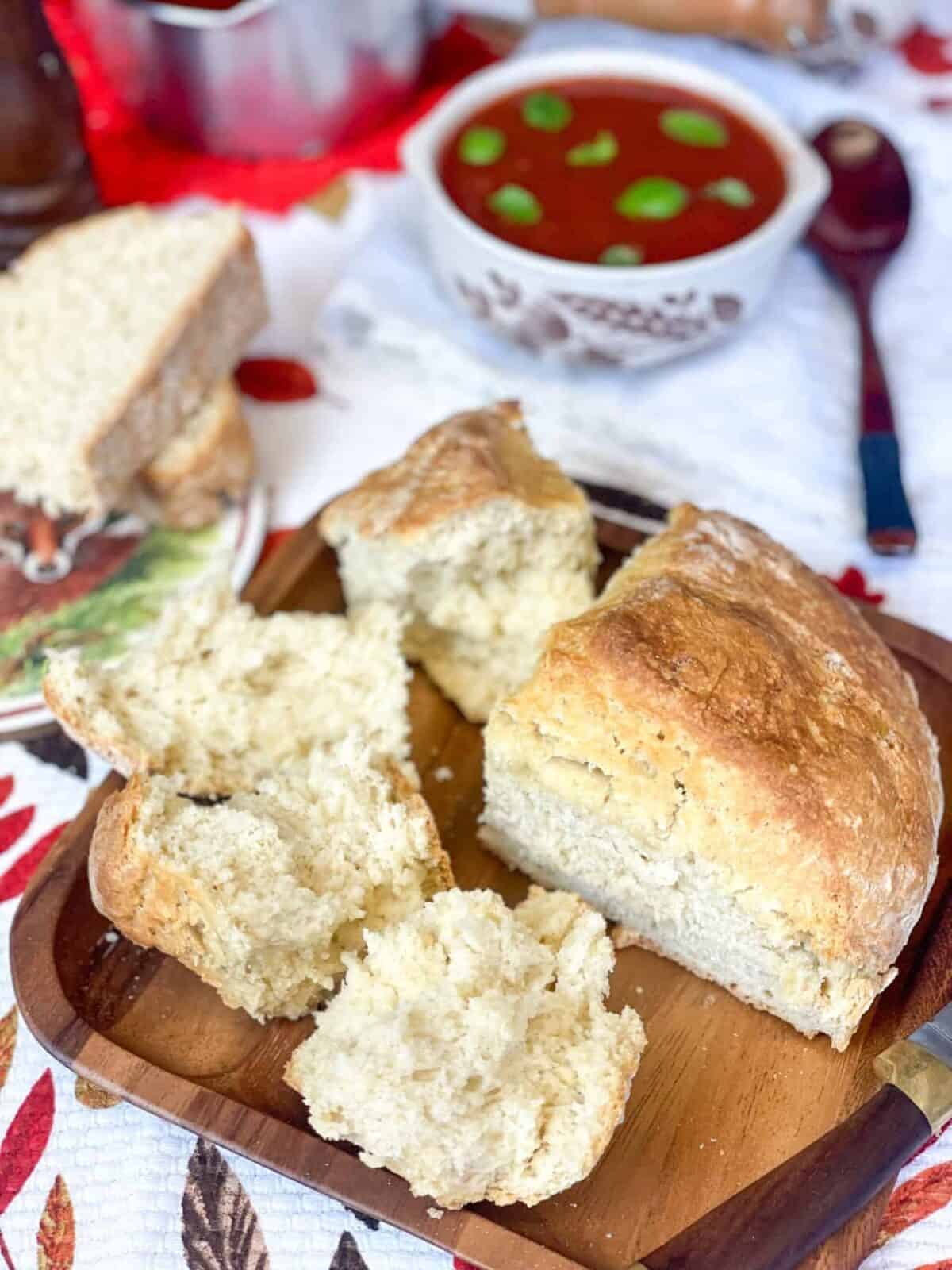 Damper bread in pieces with an intact triangle of bread, slices of bread to side, and tomato soup served in background, leaf patterned tea towel background.