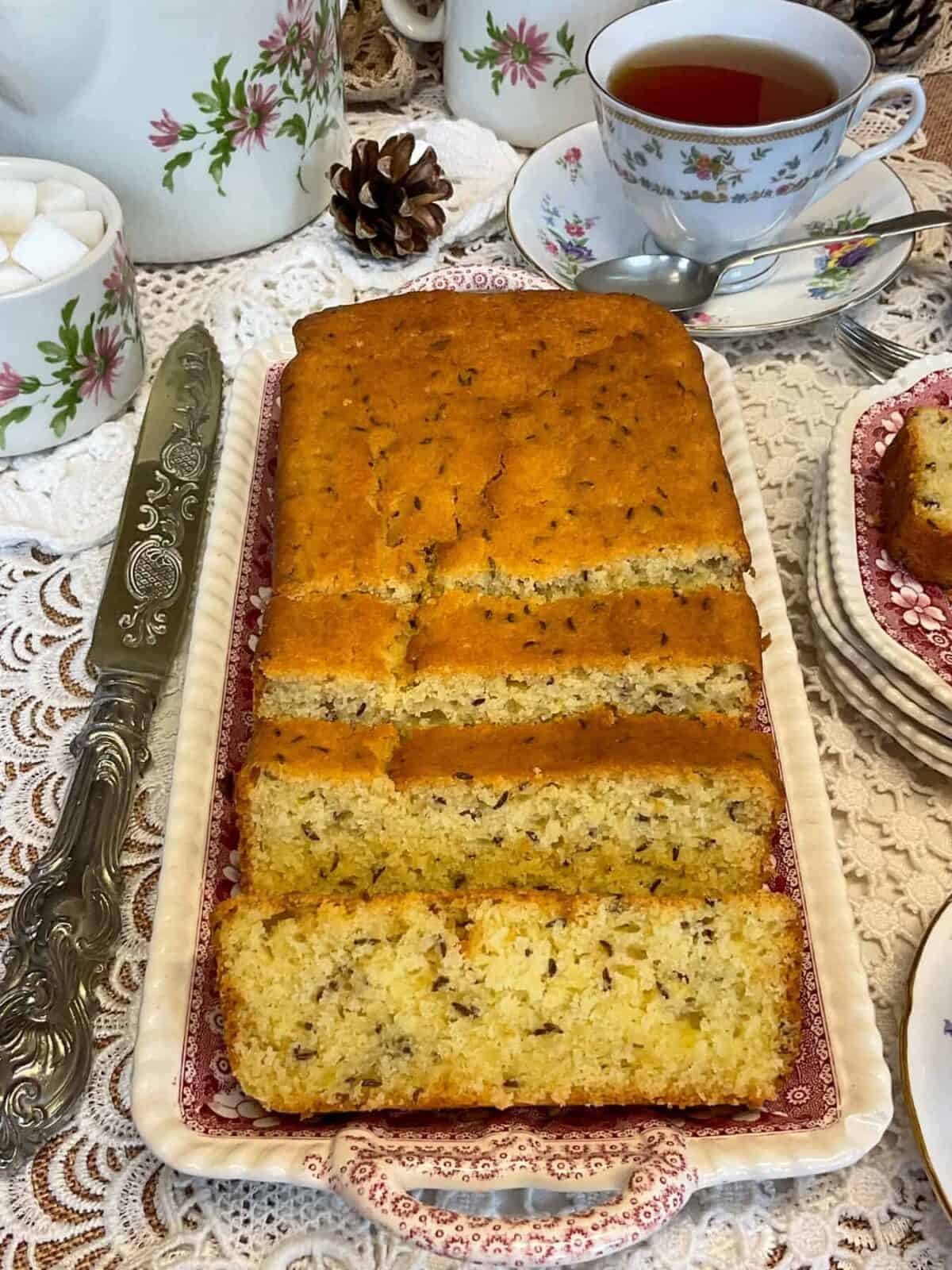 A seed cake on a serving tray with silver knife to side, tea cup and saucer, sugar bowl to side.