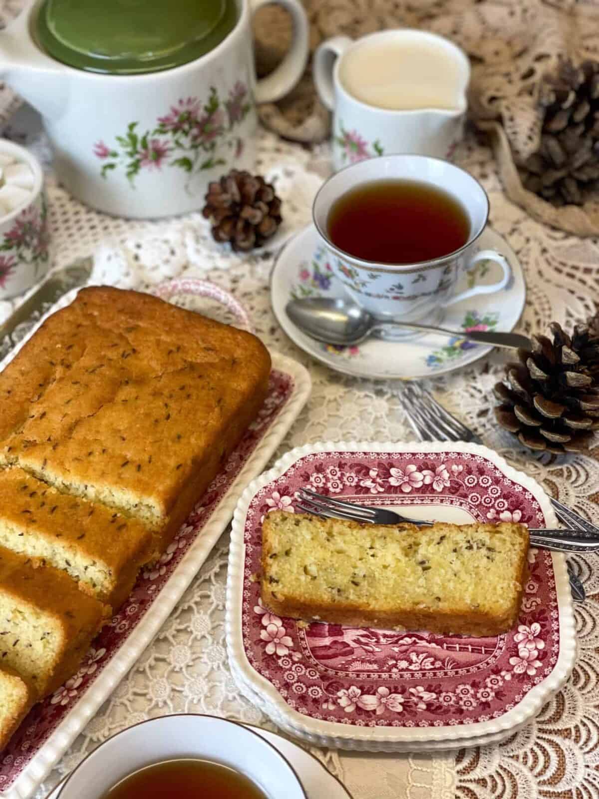 Tea table set with slice of seed cake and vintage tea cups and saucers, green lidded teapot with flower image in background, with cake tray to side.