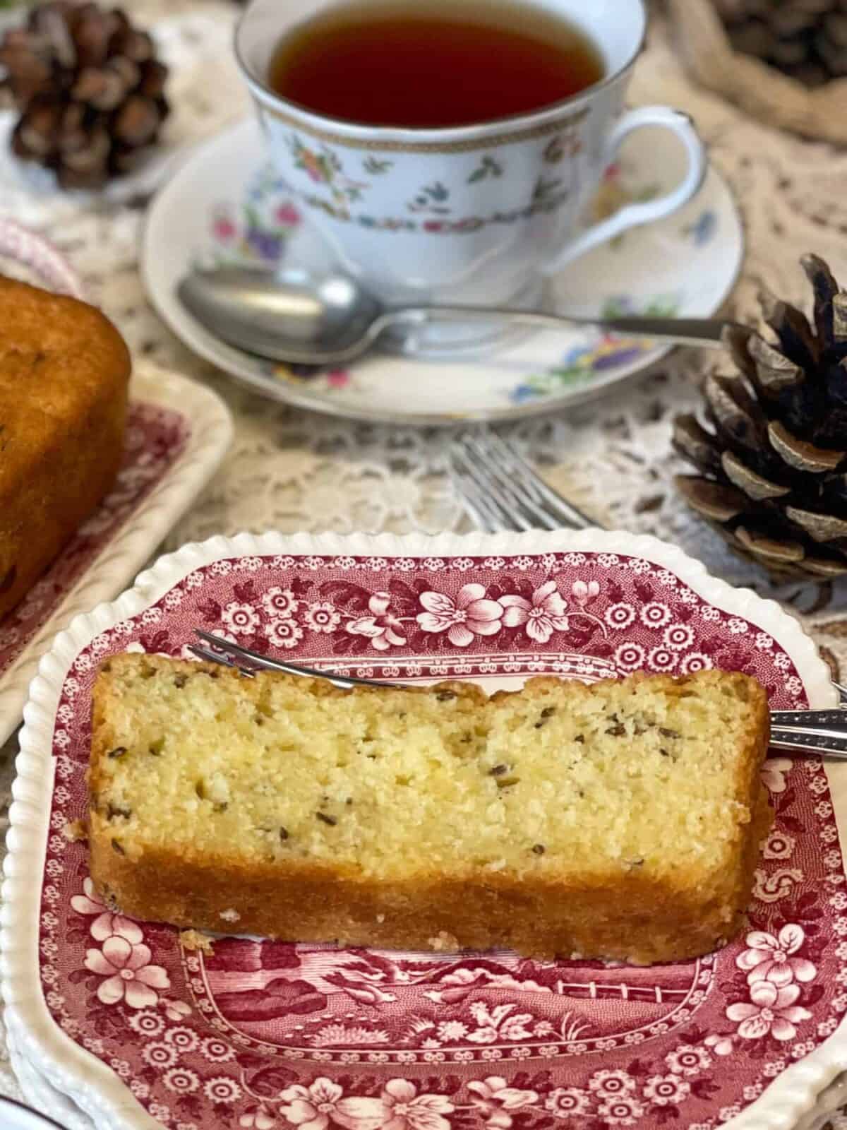 A slice of seed cake on a red patterned vintage small plate with tea cup and saucer with spoon in background.