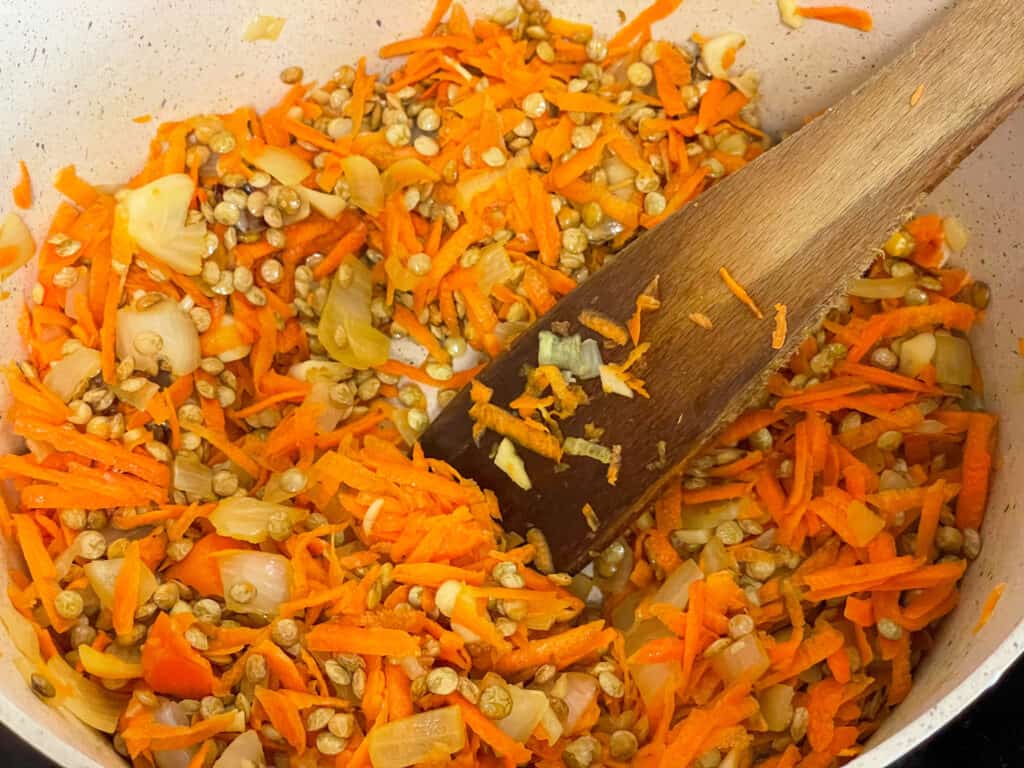 Grated carrot and green lentil in pan with onions with wooden spatula.