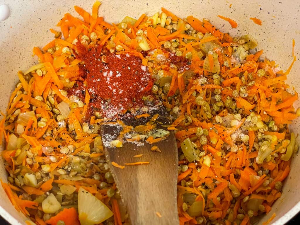 Paprika added to cooking lentils, carrot and onion in white pot with wooden spatula.