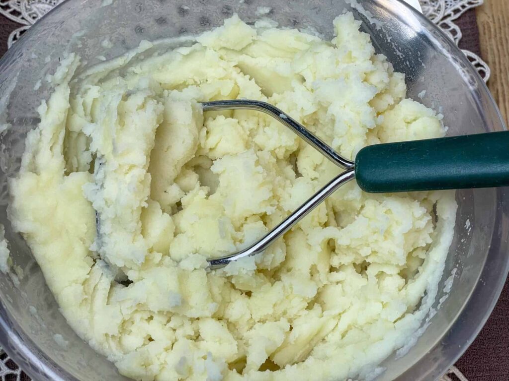 Mashed potato in plastic bowl with potato masher with green handle.