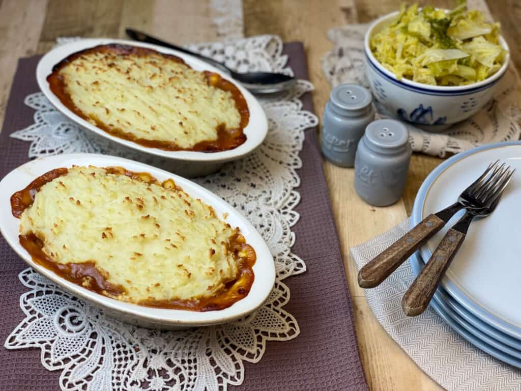 Two white oval pie dishes sitting on white rectangle doily and brown tea towel, serving plates to side along with salt and pepper shakers, and bowl of cabbage, forks to side, wooden table background.