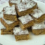 Close up of coconut, cornflake and chocolate crunch traybake sitting in a pile on top of a white plate, white doily background, featured image.