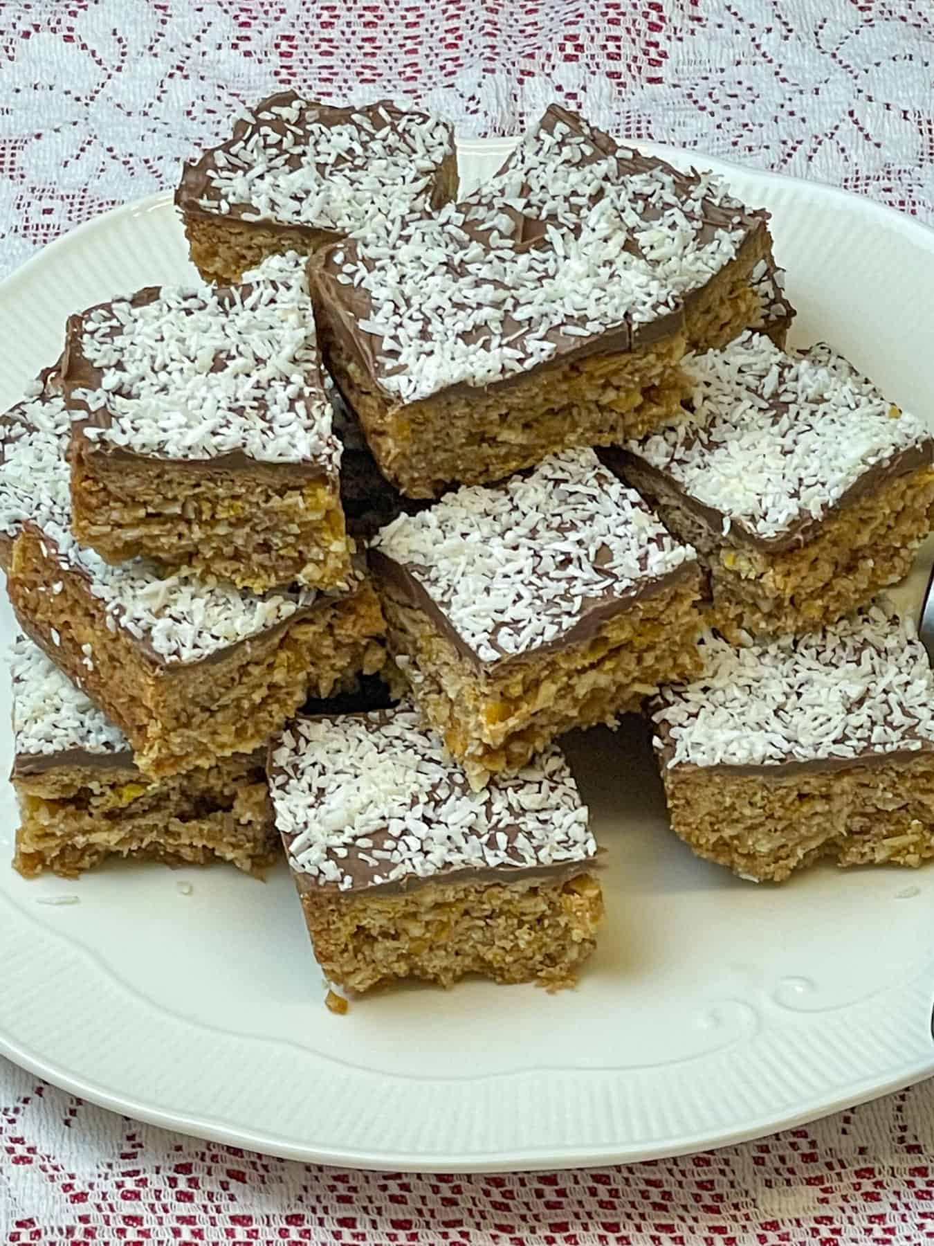 Close up of coconut, cornflake and chocolate crunch traybake sitting in a pile on top of a white plate, white doily background, featured image.