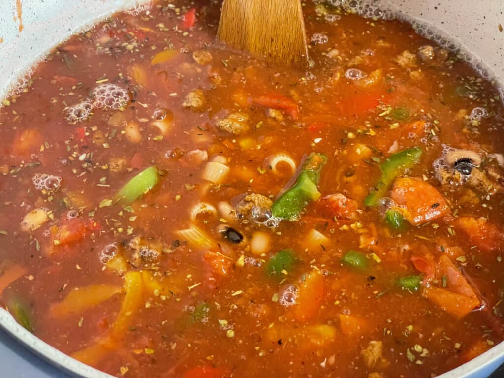 vegetable stock added to the American goulash ingredients within the soup pan, wooden spatula stirring the mixture.
