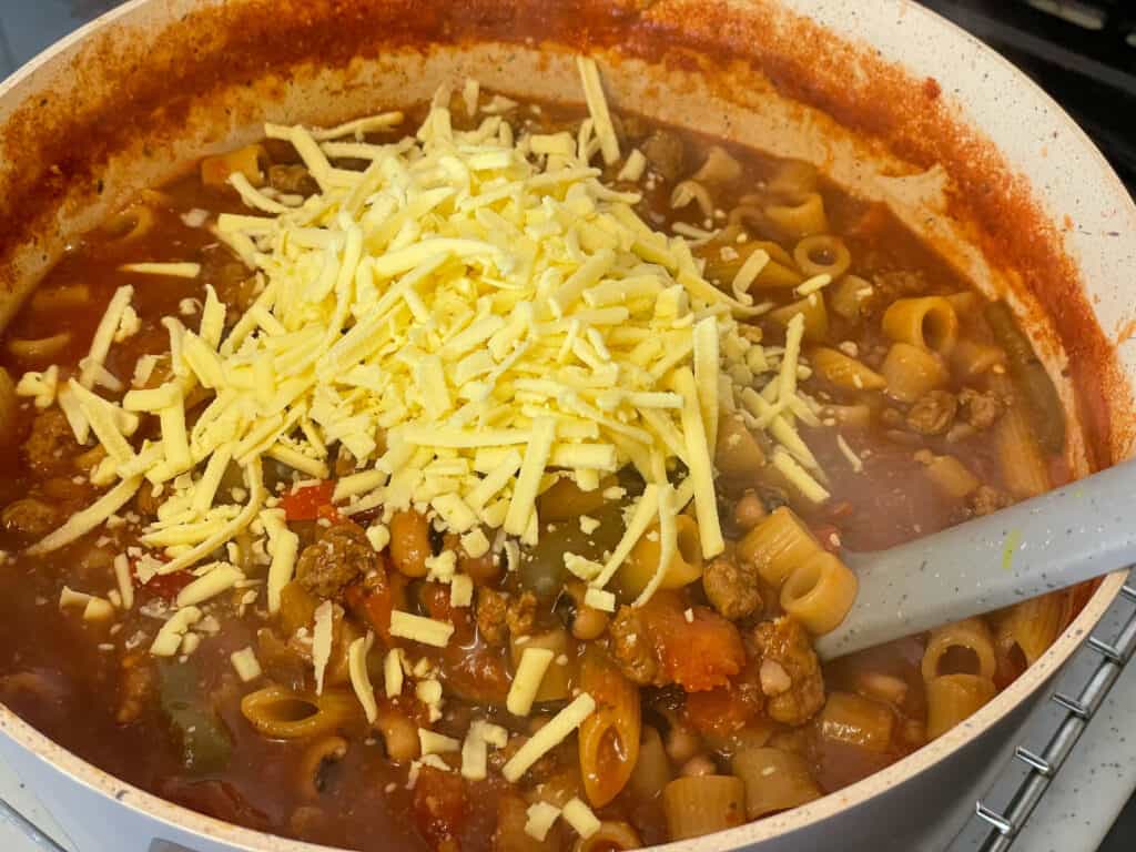 grated vegan cheese added to American goulash in soup pan with grey ladle.