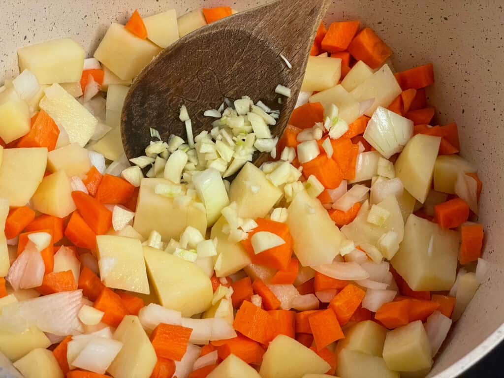 Diced potato, carrot, onion and garlic added to white pan with wooden spoon.