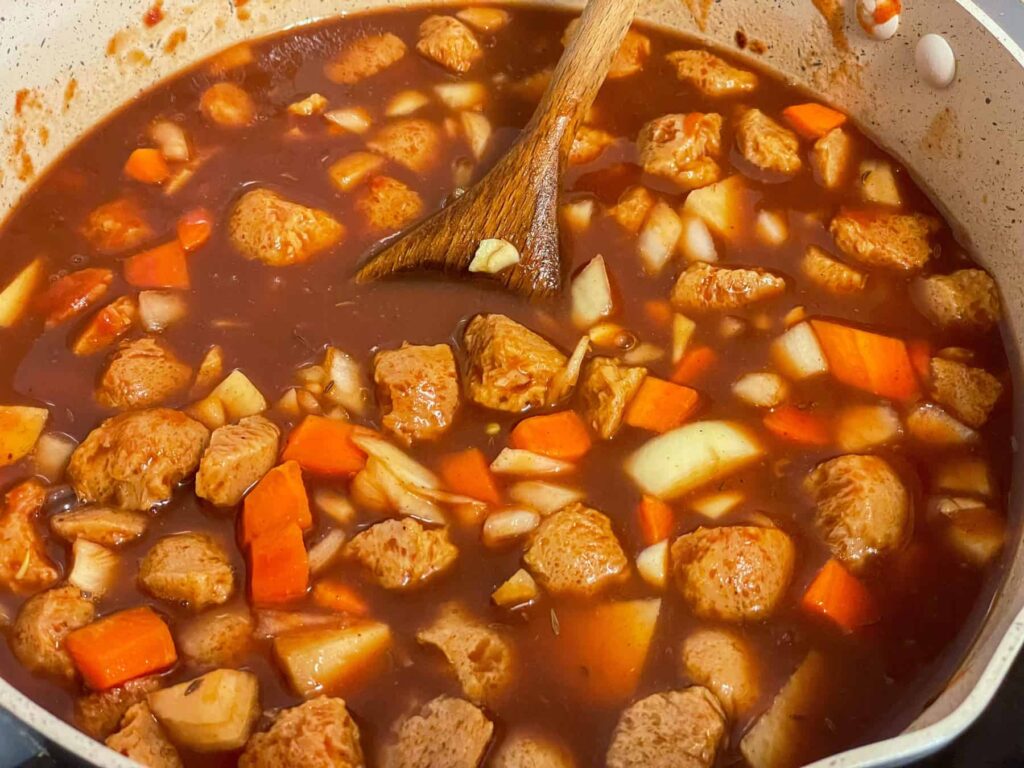 Veggie stock poured into the pan with veggies for Hungarian goulash, wooden spoon to side.