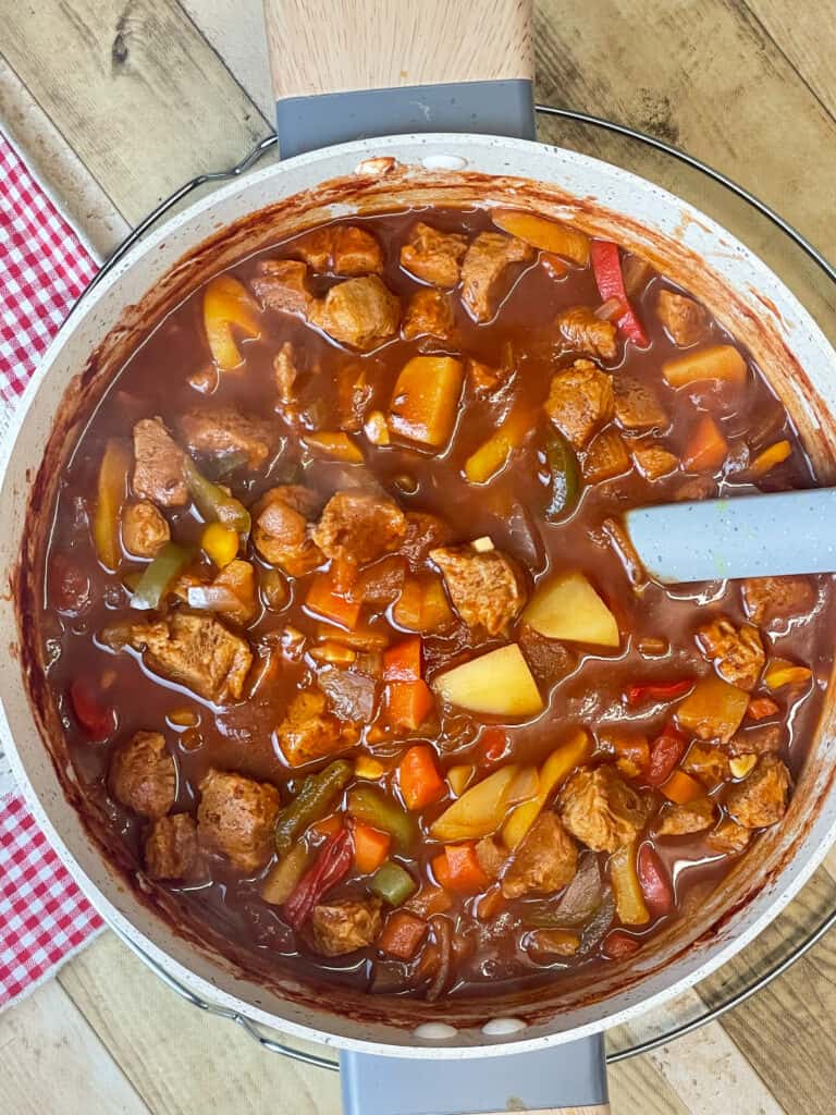 Vegan Hungarian goulash in large pot with wooden handles, wooden background and red check placemat to side, grey ladle in pot.