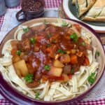 A red clay dish full of thick pasta noodles and Hungarian goulash stew on top with a sprinkle of parsley, small brown cup to side with paprika flakes and green plate with garlic bread slices, red and white check placemat, featured image.