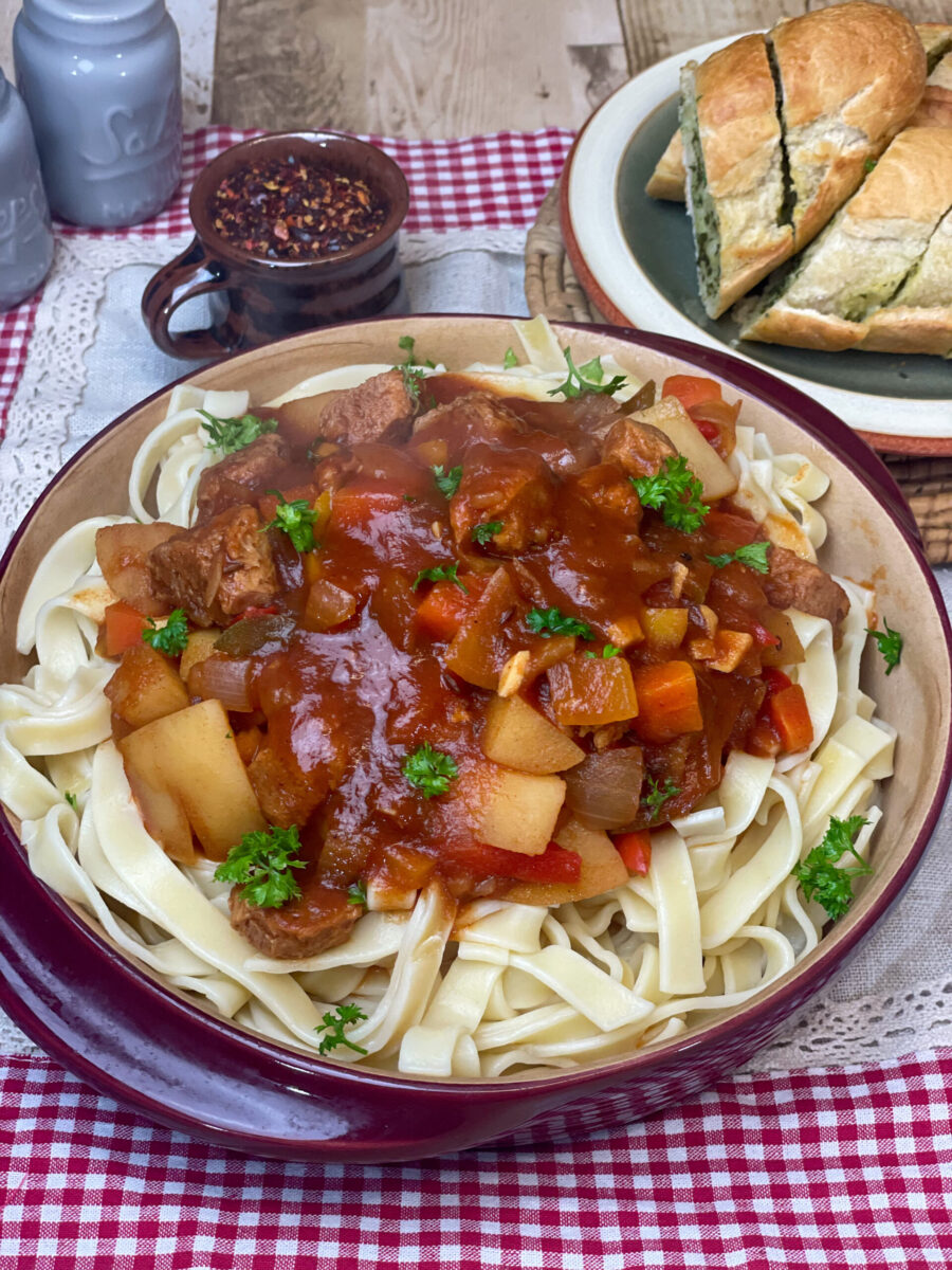 A red clay dish full of thick pasta noodles and Hungarian goulash stew on top with a sprinkle of parsley, small brown cup to side with paprika flakes and green plate with garlic bread slices, red and white check placemat, featured image.