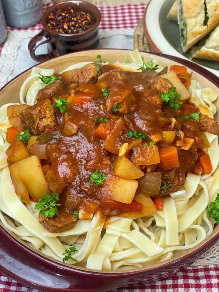 close up of vegan Hungarian goulash with parsley sprinkled over, small mug with paprika flakes and garlic bread to side.