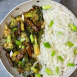 vegan Mongolian beef and broccoli cooked in grey and white wok with rice noodles and spring onion garnish, grey background, featured image.