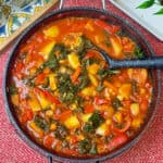 skillet pan full of Spanish chickpea stew with ladle, red background with bread tray to side with tomato bread and olive oil jug to side, featured image.