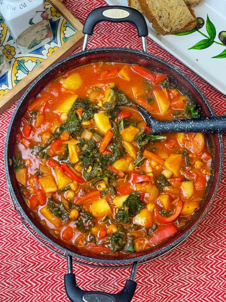 skillet pan full of Spanish chickpea stew with ladle, red background with bread tray to side with tomato bread and olive oil jug to side, featured image.