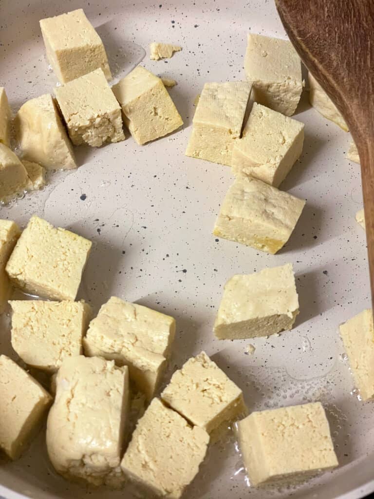 tofu cubes added to cream coloured pan with wooden spatula to side.