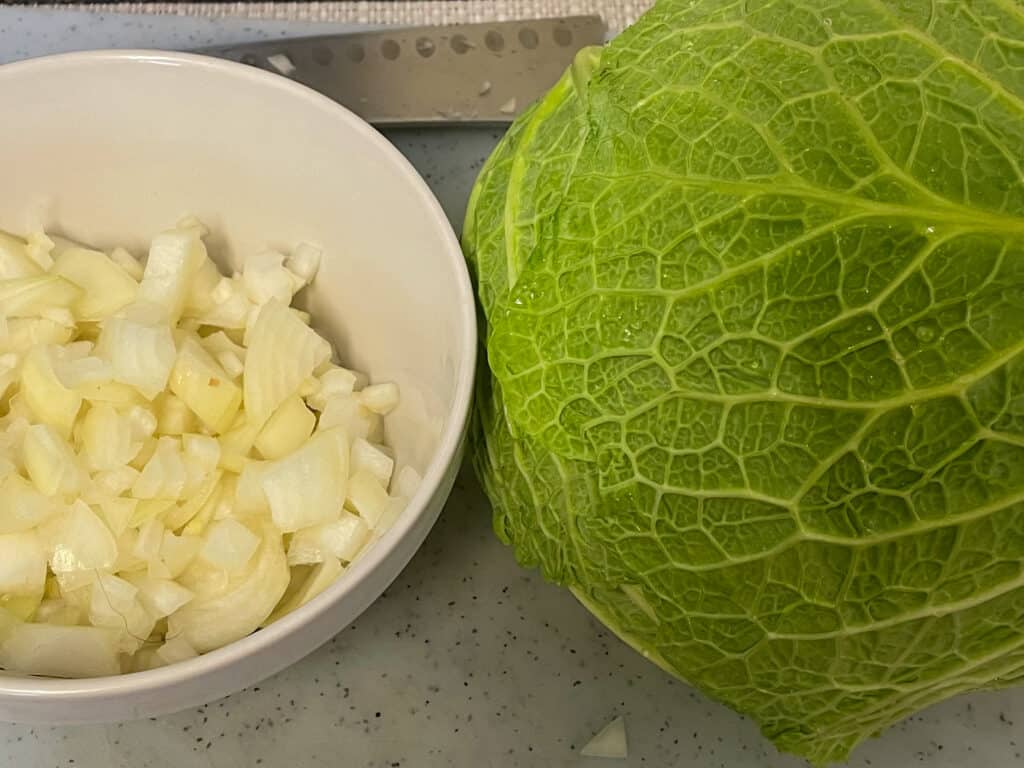 a whole head of cabbage on white chopping board with small dish of chopped onion.