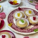 red patterned plate with four British vegan fairy cakes and kola sweeties, roses on white tablecloth, small dish of sweets, small red plate with one fairy cake, teapot to back, featured image.