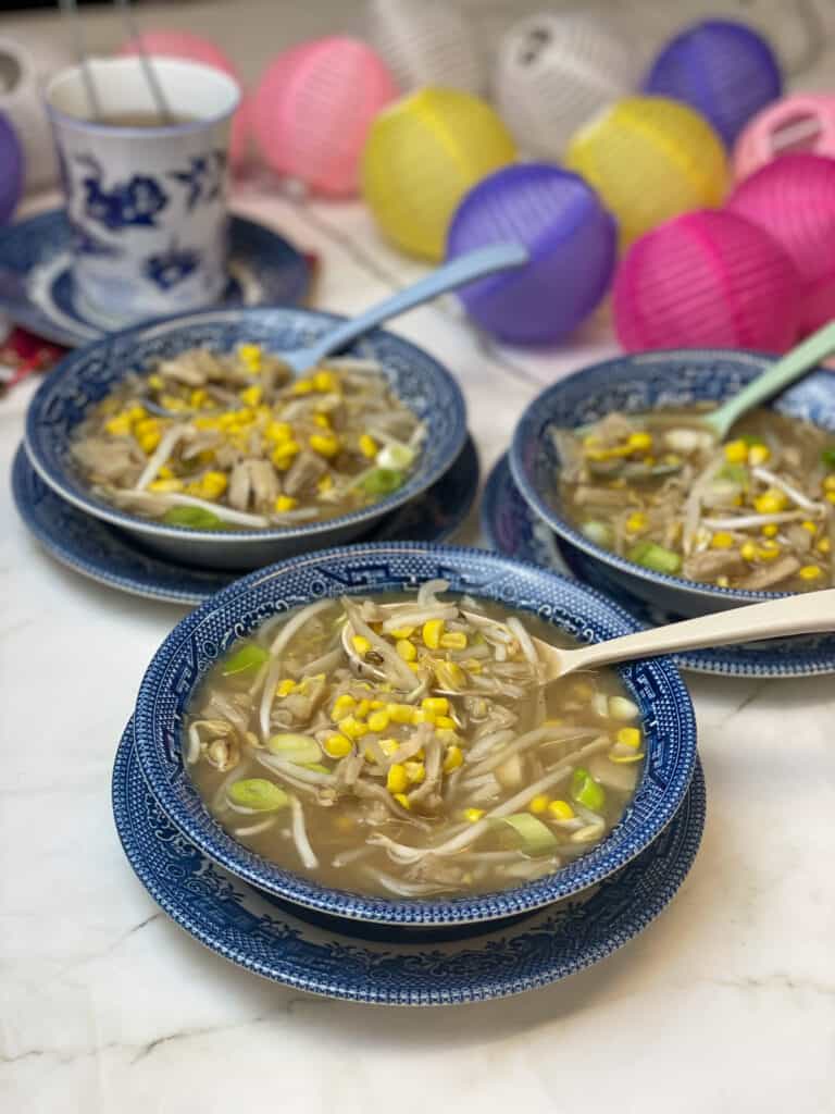 Chinese sweetcorn soup served in three blue patterned bowls with blue plates under each bowl, colourful paper light balls to side decor and Chinese cup to side with saucer.
