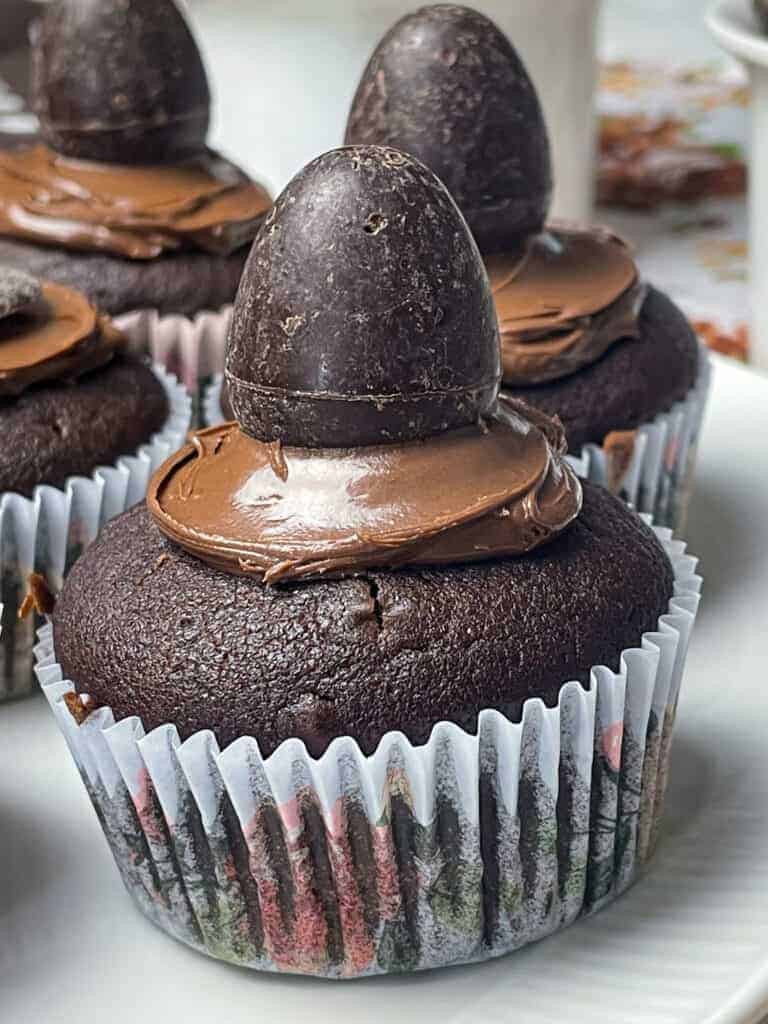 close up of eggless chocolate bun with icing and chocolate egg decoration with three buns in distance, white plate background.