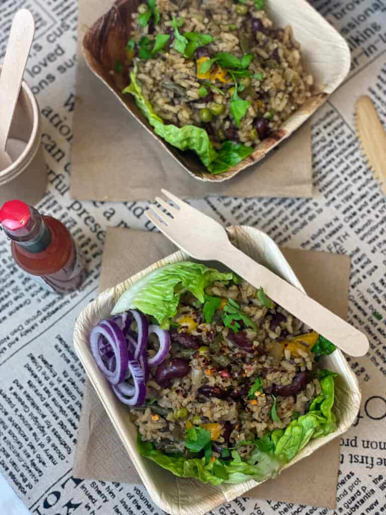 two take-out containers with Cajun dirty rice on tan napkins and newspaper print background, bottle of hot sauce to side.