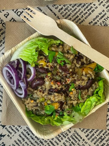 vegan Cajun dirty rice in a bamboo take-out container with wooden fork, tan coloured napkin underneath and newspaper print table mat, featured image.