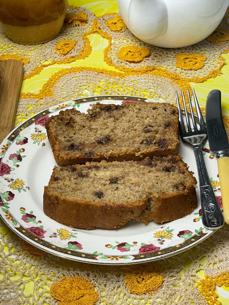 Two slices of banana bread served on small flower patterned plate with cake fork and knife, yellow net place mat, white teapot to side and yellow mug to side, yellow tablecloth background.