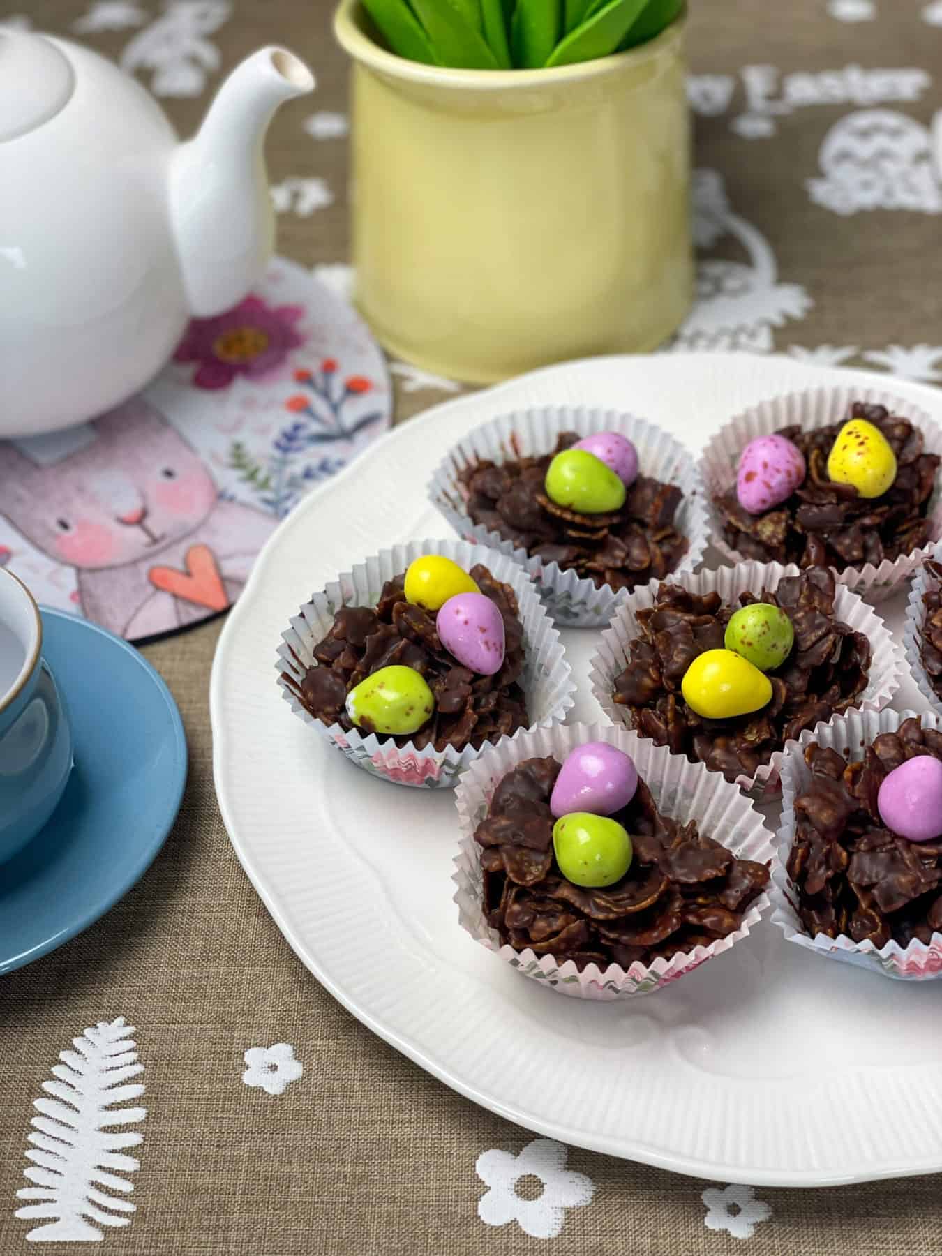 A large white plate full of chocolate cornflake cakes, white tea pot to side, blue tea cup and saucer to side, yellow jug with flower stems to background, rabbit image tea pot placemat.