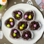 Vegan Easter Cornflake Cakes on a white plate with vegan mini eggs on top of each cake and a linen table runner with writing stating 'happy Easter', rabbit placemat image and peach tulips to side/featured image.