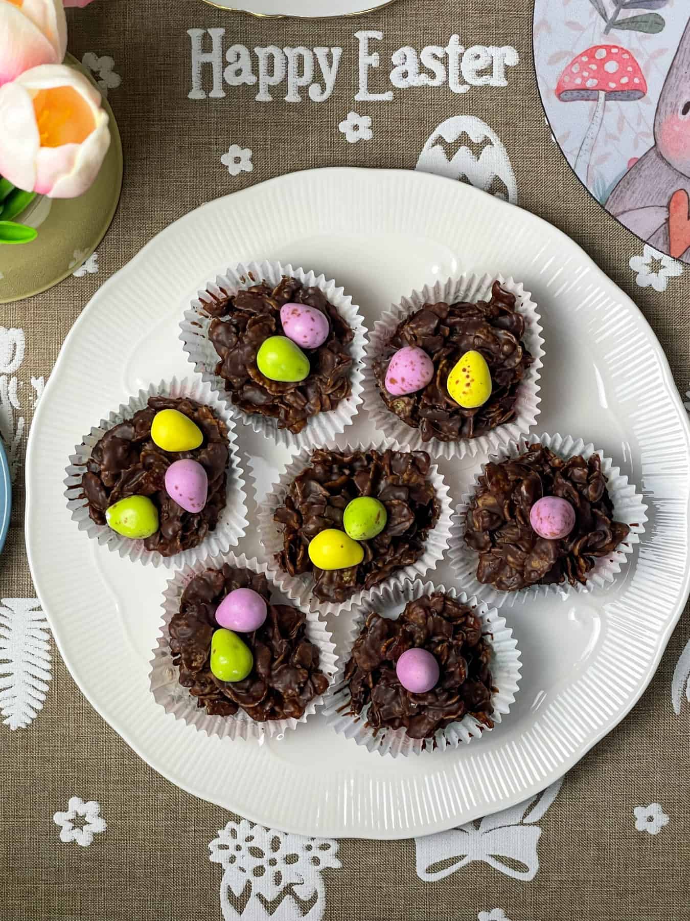 Vegan Easter Cornflake Cakes on a white plate with vegan mini eggs on top of each cake and a linen table runner with writing stating 'happy Easter', rabbit placemat image and peach tulips to side/featured image.