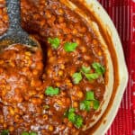 featured image for baked beans in tomato sauce, side view of pan full of beans garnished with parsley and ladle in pan, red background.