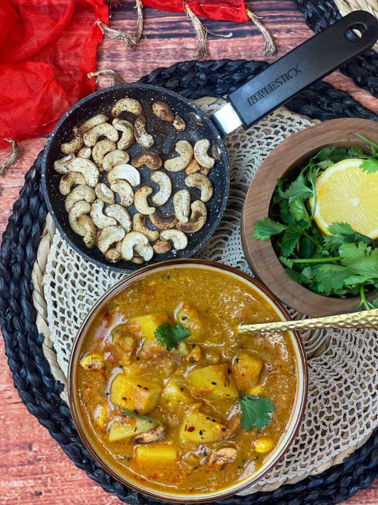 Bombay potato curry soup served with small pan of toasted cashew nuts and wooden mortar bowl with fresh cilantro and slice of lemon in bowl, mat background with orange scarf to side, Featured Image.