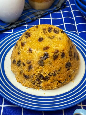 spotted dick pudding on a blue rimmed white serving plate with blue and white check table cloth, custard and cream jugs to side, Featured Image.
