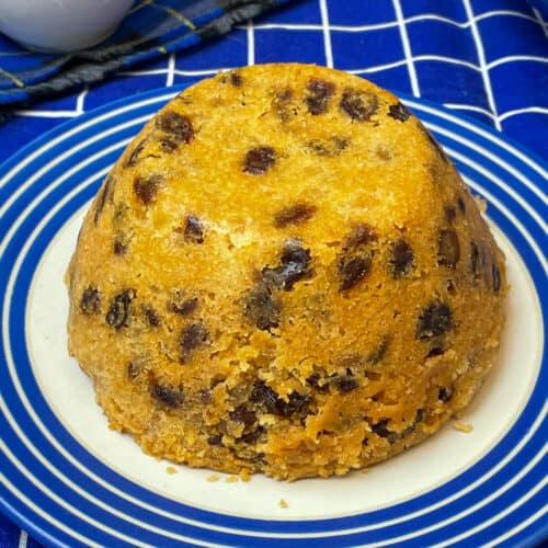 spotted dick pudding on a blue rimmed white serving plate with blue and white check table cloth, custard and cream jugs to side, Featured Image.