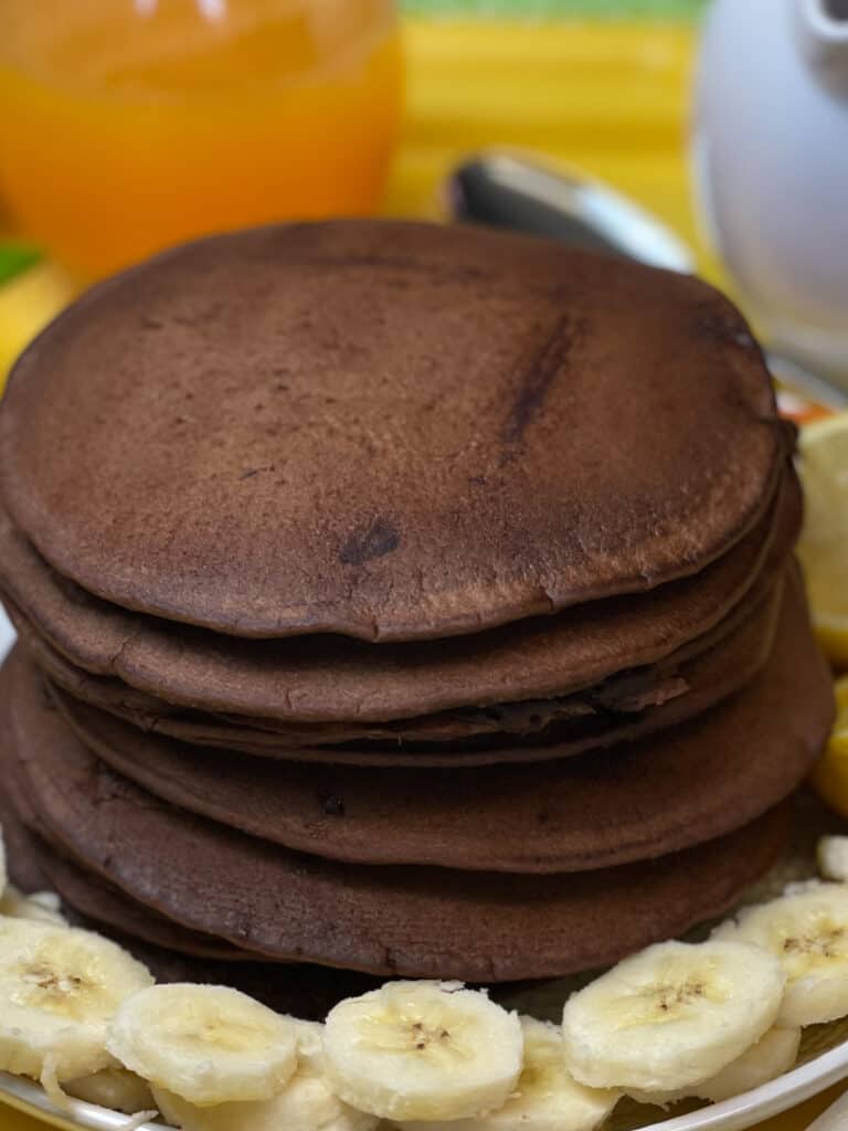 stack of cooked chocolate chip pancakes on serving plate with sliced bananas, glass of orange juice to side and syrup jug to back, on yellow tray.