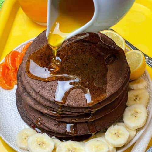 vegan chocolate chip pancakes with a jug drizzling syrup over the pancake stack, sliced bananas and orange segments around the serving plate, glass of orange juice to side, yellow serving tray, featured image.