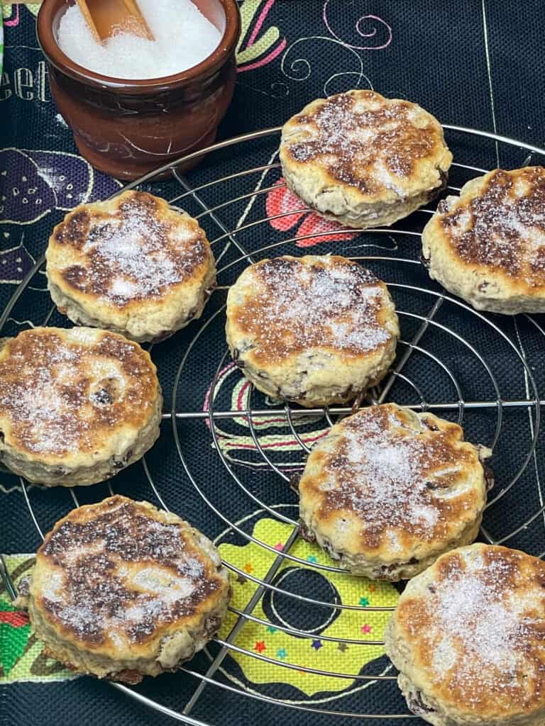 vegan Welsh cakes on wire rack cooling with a sprinkling of white sugar over each cake, small brown sugar bowl to side with small wooden shovel, black tea towel background.