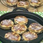 Vegan Welsh cakes on a cast iron griddle with green stripy white tea towel underneath, in distance wire rack with cooked cakes and small brown sugar dish/featured image.