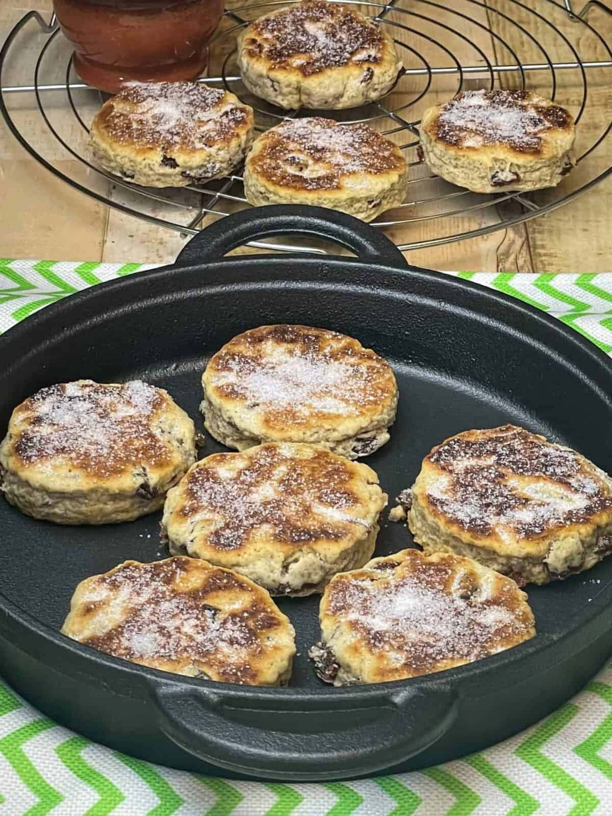 Vegan Welsh cakes on a cast iron griddle with green stripy white tea towel underneath, in distance wire rack with cooked cakes and small brown sugar dish/featured image.