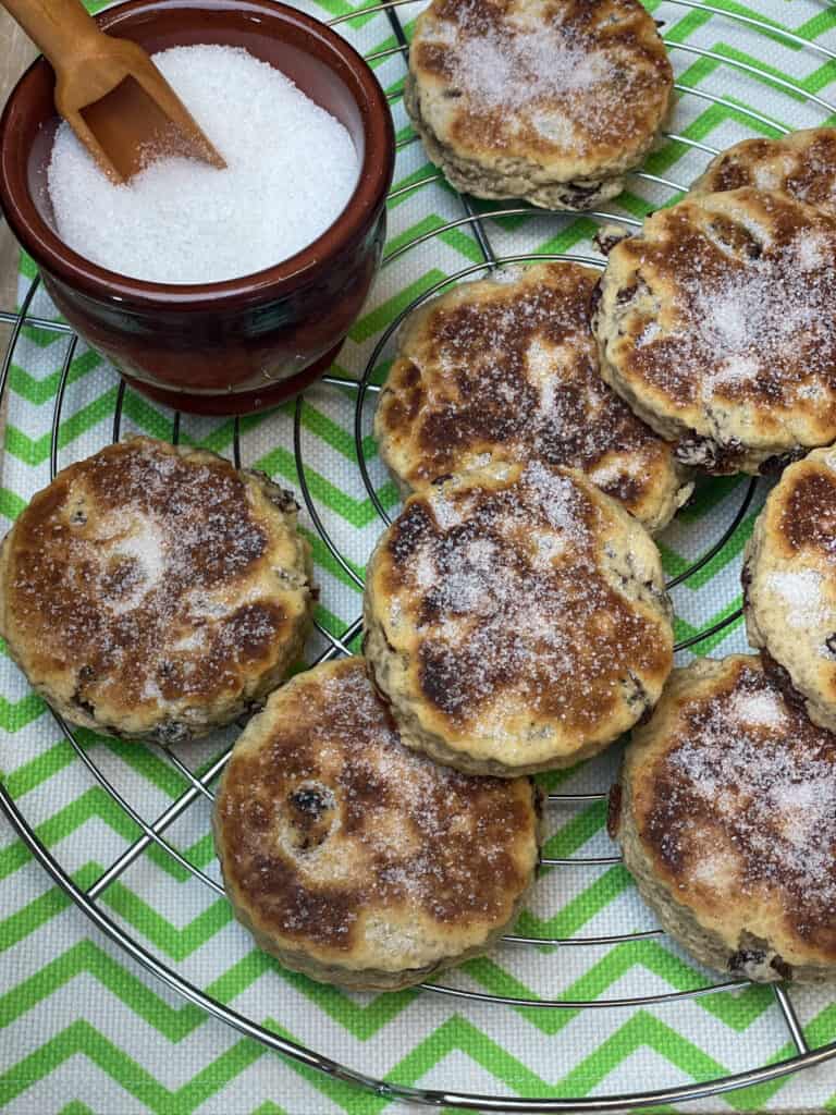 Welsh cakes on wire rack with green zig zag and white tea towel underneath, small brown sugar dish with small wooden scoop to side.