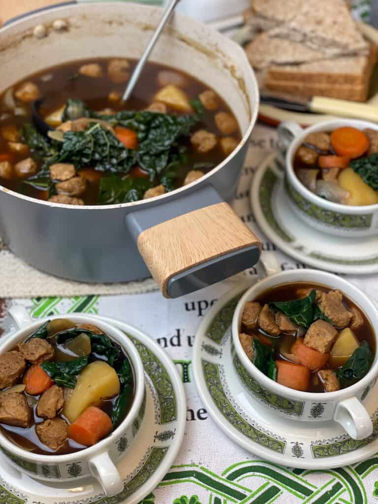 stew pan full of vegan Irish stew with ladle set out on dinner table with three small serving bowls filled with stew and plate of bread to side, with butter dish and knife, Irish tea towel background.