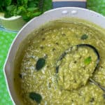 A pan full of British mushy peas with mint leaf garnish, ladle in pan, yellow jug of mint stalks to side, and green background, featured image.