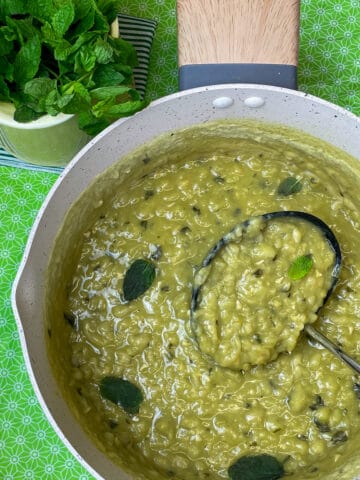 A pan full of British mushy peas with mint leaf garnish, ladle in pan, yellow jug of mint stalks to side, and green background, featured image.