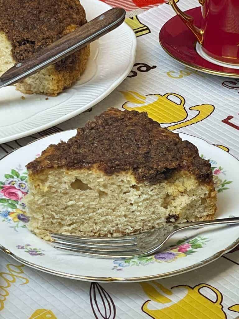 slice of coffee cake on flower patterned cake plate and cake fork, red coffee mug to side with whole cake at edge of photo with cake slice handle.