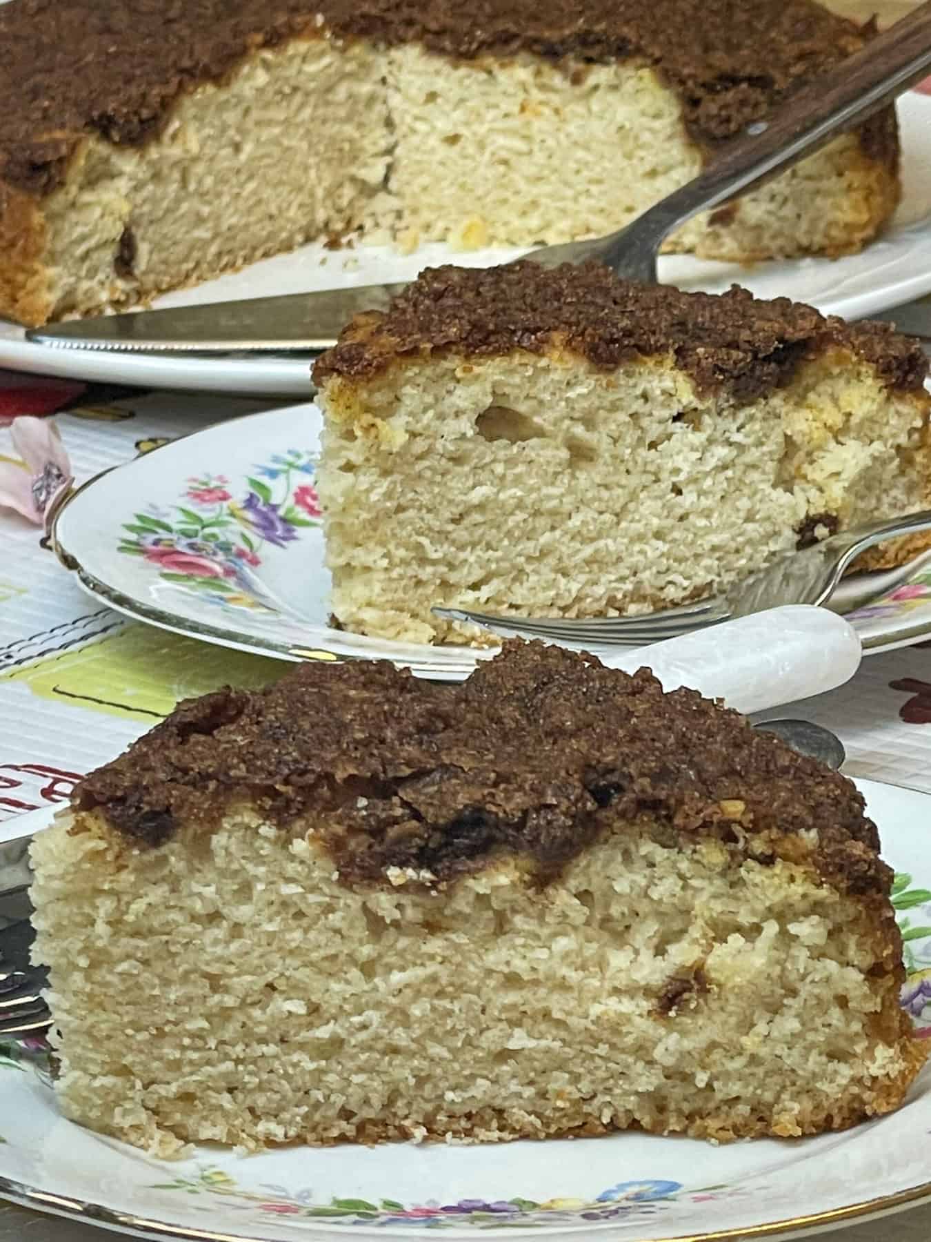 two slices of cinnamon streusel topped cake on small flower patterned plates, whole cake in background with cake slice.